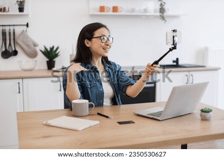 Joyful asian female in glasses holding selfie stick with smartphone while taking seat at wooden table in kitchen. Happy young woman creating self-portrait using smart gadgets in home interior.