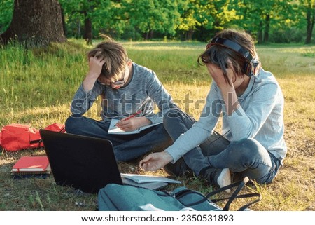 Two tired boys sitting on ground in park in front of laptop, doing homework with pencils together. Pupil in headphones and classmate in glasses weary of writing assignment in notebooks. Royalty-Free Stock Photo #2350531893