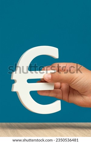 White Euro symbol in a hand on a blue background close-up with copy space. Euro exchange rate, economy in Europe or inflation.