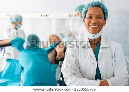 Portrait of successful smiling African American female doctor gynecologist with surgical cap and uniform looking at camera standing in front of medical team and woman in labor. Royalty-Free Stock Photo #2350555795