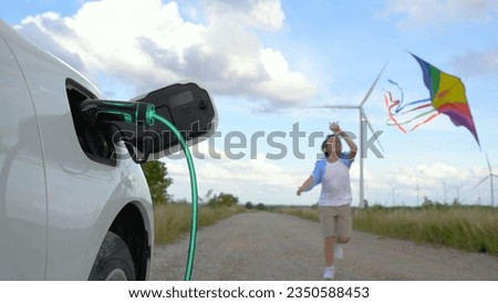 Focus eco-friendly EV car recharging battery from charging station using renewable and clean wind turbine generator with happy young boy running in the road and playing with kite in background. Peruse