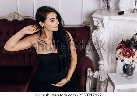 Pregnant woman strokes hugging the belly tummy abdomen enjoying pregnancy sitting on maroon couch dressed black suit. Future family, baby infant expecting child inside
