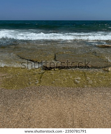 photo of big rocks and wavy sea on the beach on a hot sunny day in summer. Blue sky, big tall boulders on mossy sea shore with sea water.
