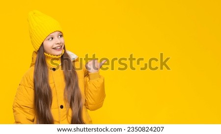 A happy little girl in an autumn jacket and hat points to your advertisement and smiles broadly. Yellow isolated background. Copy space. Banner.