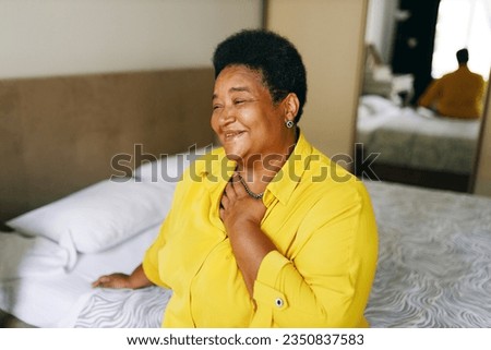 Happy cheerful grateful african american grandmother sitting on bed in yellow shirt keeping hand on chest smiling, being thankful for best life, thinking about her past, funny unforgettable moments Royalty-Free Stock Photo #2350837583