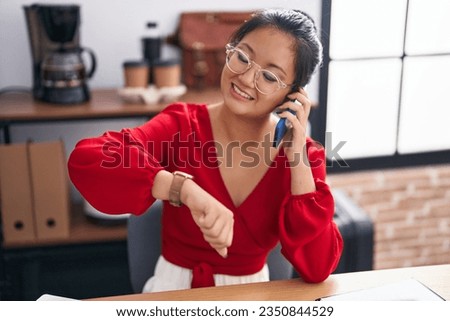 Young chinese woman business worker talking on smartphone looking watch at office