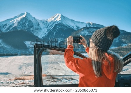 Woman taking pictures of winter mountain view mobile phone, standing near the car. Woman travel exploring, enjoying the view of the mountains, landscape, lifestyle concept winter vacation