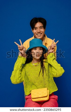 Positive young asian couple wearing colorful clothes showing peace sign while posing isolated over blue studio wall