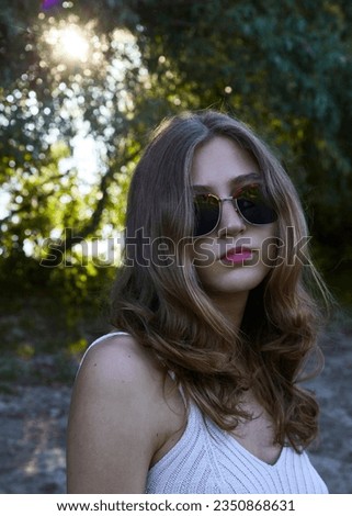 a beautiful woman in sunglasses poses looking at the camera while walking in the park on a sunny day