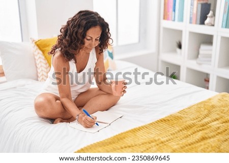 Middle age woman drinking cup of coffee writing on notebook at bedroom