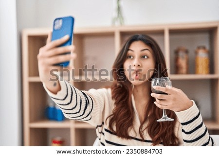 Hispanic young business woman taking a selfie picture drinking a glass of wine making fish face with mouth and squinting eyes, crazy and comical. 