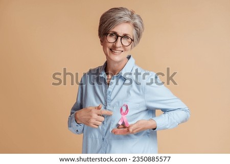 Smiling senior woman wearing eyeglasses showing pink ribbon isolated on beige background. Health care, support, prevention. Breast cancer awareness month concept Royalty-Free Stock Photo #2350885577