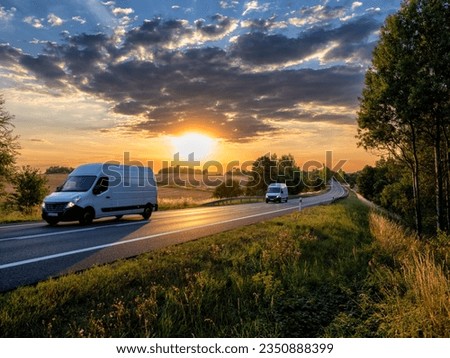 Two white fast delivery vans driving on the asphalt road in rural landscape at sunset with dark cloud Royalty-Free Stock Photo #2350888399