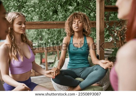 smiling african american woman meditating in lotus pose and holding hands with girlfriends in patio