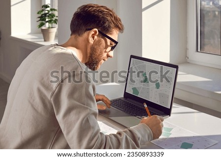 Cadastral engineer keeps records of land plots by making marks on map. Young engineer with cadastral plans and projects is sitting at desk with laptop in office. Real estate object, ownership rights. Royalty-Free Stock Photo #2350893339