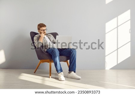 Pensive smart clever young man coder in casual shirt, jeans and glasses sitting in comfortable armchair by light gray wall, using modern laptop computer, working on website, thinking with hand on chin Royalty-Free Stock Photo #2350893373