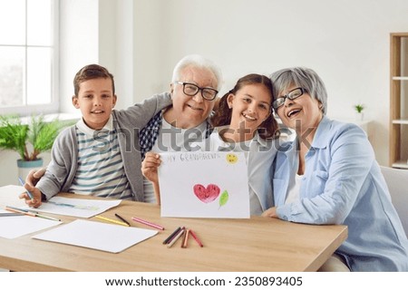 Happy grandparents and grandchildren spend time together. Indoor family portrait joyful grandma, grandpa and little children sitting at table with pictures and pencils, looking at camera and smiling