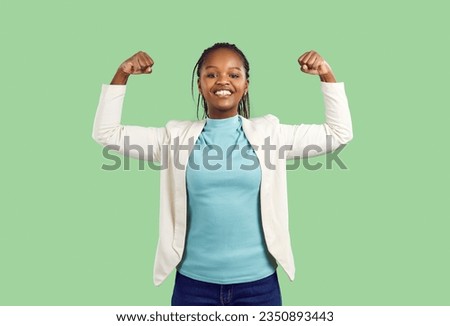 Young African woman demonstrates her strength. Happy active strong African American model standing on solid green colour studio background feeling confident and flexing both her arms. 