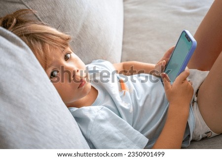 Close up cute little boy using smartphone, looking at screen, curious child holding phone in hands, sitting on couch at home alone, playing mobile device game, watching cartoons online.
