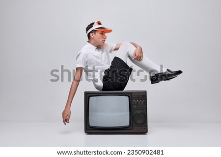 Portrait of stylish young man, boy posing in white tights and blac shirt, sitting on retro TV isolated over grey background. Concept of modern fashion, art photography, style, queer, uniqueness, ad