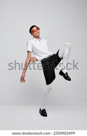 Portrait of stylish boy posing in black shorts, white shirt and tights with red glasses isolated over grey background. Concept of modern fashion, art photography, style, queer, uniqueness, ad