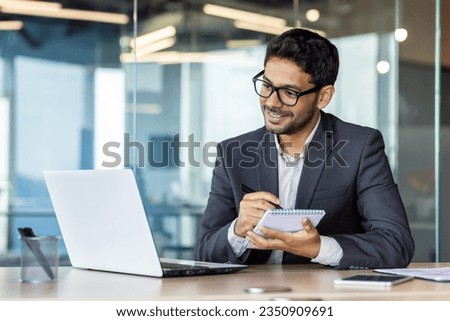 Mature experienced hispanic businessman in middle of office watching online webinar and training course, man using laptop for video call, writing data in notebook, smiling excitedly and satisfied.