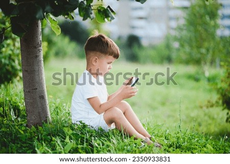The child looks into the phone while sitting in the summer on the street. Little boy with a mobile in his hands.