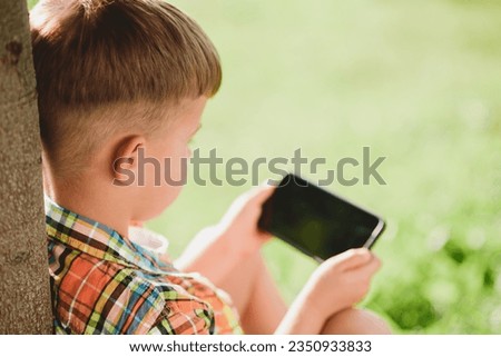 The kid looks into the mobile while sitting in the summer on the street. Small child with a phone in his hands.