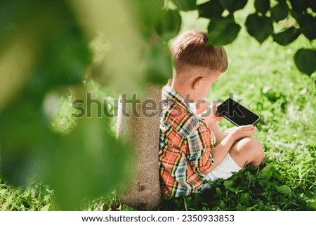 Cheerful boy sitting on the grass looks cartoons in the phone in the summer at sunset. Cute baby having fun in nature