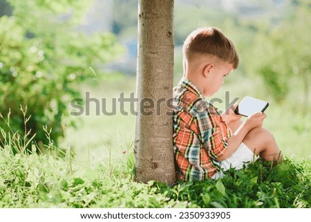 Cheerful boy sitting on the grass looks cartoons in the phone in the summer at sunset. Cute baby having fun in nature