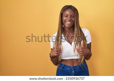 African american woman with braided hair standing over yellow background success sign doing positive gesture with hand, thumbs up smiling and happy. cheerful expression and winner gesture. 