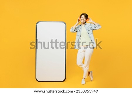 Full body young woman she wears green t-shirt denim shirt casual clothes big huge blank screen mobile cell phone smartphone with area listen music oin headphones isolated on plain yellow background