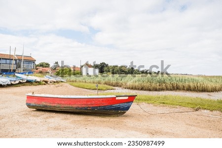 A red and blue wooden boat pictured on the shore at Brancaster Staithe on the North Norfolk coast behind its rope and anchor.