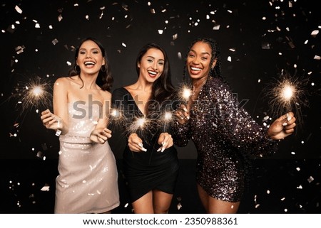 Three happy beautiful young ladies in nice dresses with bengal lights posing in confetti over black background, multiracial girlfriends celebrating New Year together, have xmas party Royalty-Free Stock Photo #2350988361