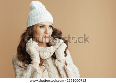 Hello winter. pensive trendy woman in beige sweater, mittens and hat on beige background.