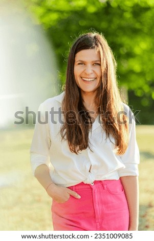 Portrait of beautiful smiling woman in white shirt looking at camera, standing in park. Breast cancer awareness month concept