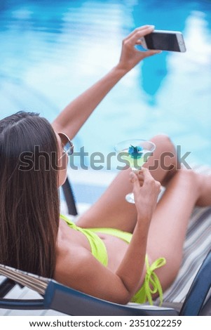 Back view of pretty girl in green swimwear and sun glasses drinking cocktail and making selfie using a phone while sunbathing on the chaise longue near the pool