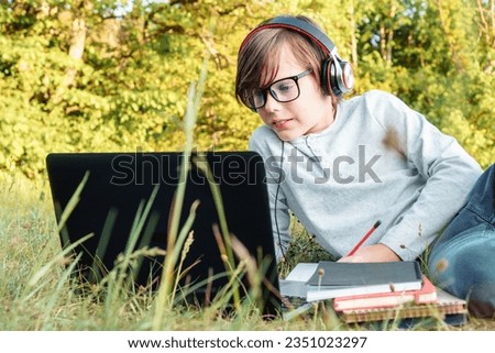 Focused junior student in glasses and earphones on head attentively watching exercises on laptop, writing task in notebook while lying on grass in park. Boy holding pencil and studying online.