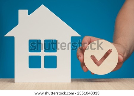 Real estate mortgage approval. Check mark icon in hand and house model as a symbol of a successful real estate purchase or sale deal. Photo with blue background and copy space.