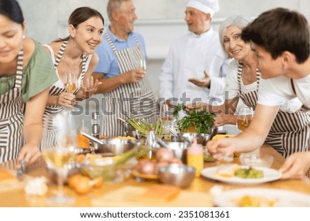 Cheerful learners of culinary classes engaged in spirited conversation around kitchen table while learning cooking fish Royalty-Free Stock Photo #2351081361