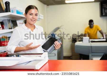 Portrait of happy young female specialist holding printed samples of notebooks in the print shop