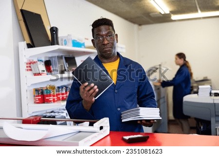 Portrait of serious middle-aged African American male specialist in uniform checking printed samples of notebooks in printer shop