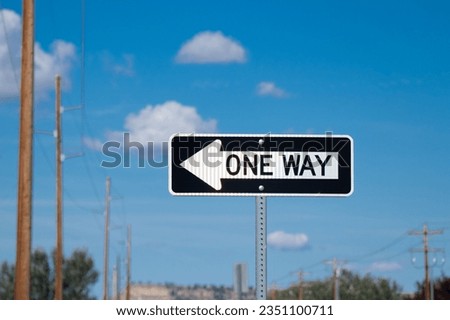 A black one way sign with a white arrow pointing drivers in the right direction; the sign is against a blue sky with white clouds. 