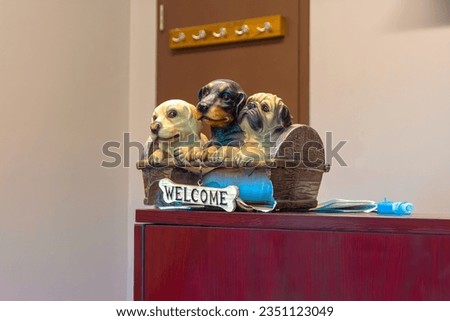 Figurine of 3 different dogs inside a basket with a welcome sign on front. The figurine is broken, is placed on top of a desk with a closed door in the background that has coat hangers.