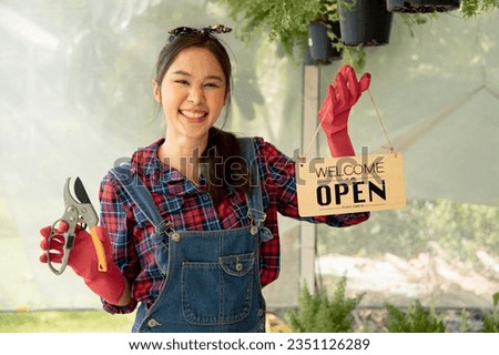 Gardener farmer standing in ornamental plants shop with text “welcome we are open”. Happy woman owner showing open sign in her small business shop. Your welcome to my small business