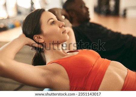 Side view of young fit female athlete in activewear doing abs crunches with hands behind head during fitness workout with black friend in gym Royalty-Free Stock Photo #2351143017