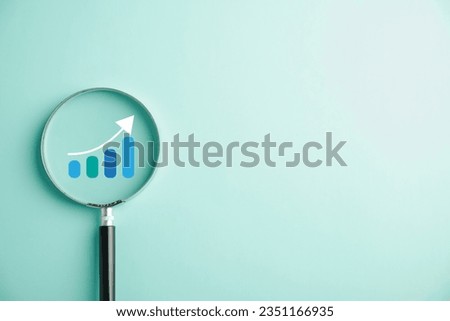 Trader hand wields magnifier glass for scrutinizing bar graph with ascending arrow. Analyzing technical stock market chart embodies the concept of business investment for potential growth and success. Royalty-Free Stock Photo #2351166935