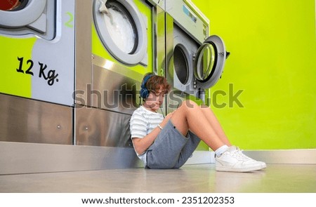 Teenage boy sitting on the floor listening to music and using his smartphone. She waits for her clothes to be washed at a self-service laundromat.