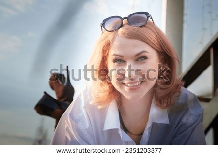 Portrait of Beautiful girl with red hair in white shirt in open wooden pavillion in village or small town. Young slender woman and sky with clouds on background on an autumn, spring or summer evening