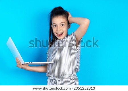 Photo of amazed Beautiful caucasian kid girl standing over blue studio background holding modern gadget arm head